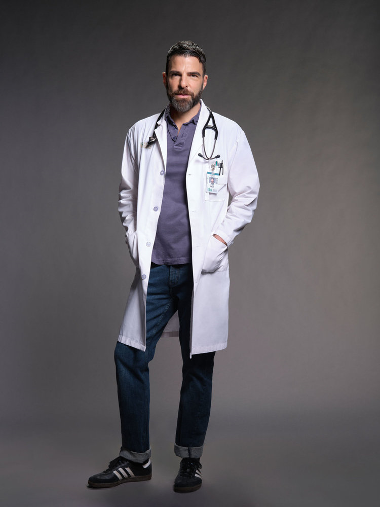 A man in a white coat and stethoscope stands confidently against a gray background, wearing a blue shirt and jeans with an ID badge.