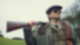 Man in a green coat and hat holds a shotgun over his shoulder. He wears sunglasses and a bandolier. Background is a green field and trees.