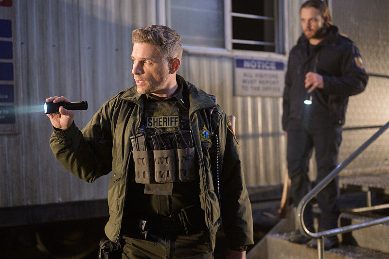 Two sheriffs in uniforms holding flashlights stand outside a building at night. One sheriff is on stairs. A sign reads "MOTEL." The mood is tense.