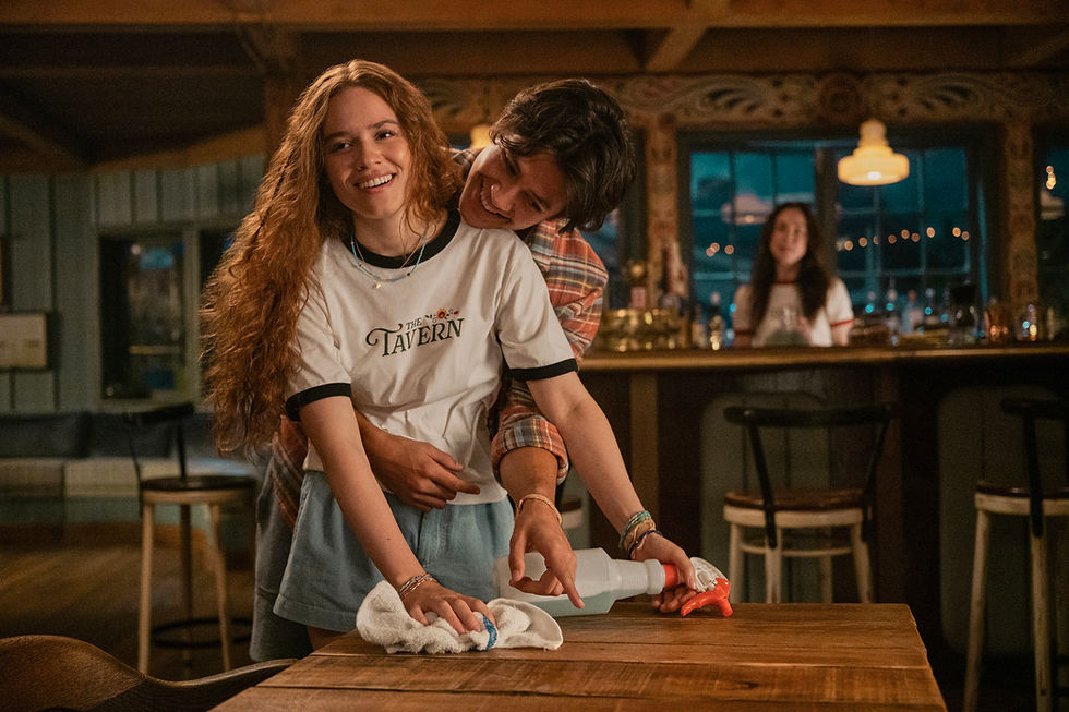 Smiling couple wipes a wooden table in a cozy tavern; her shirt says The Tavern, with warm lights and a blurred woman in back.