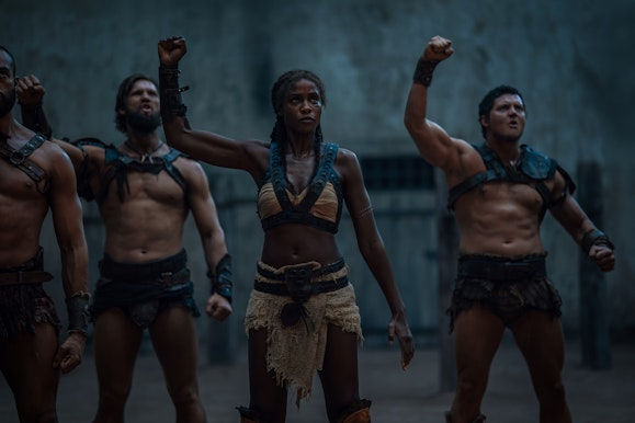 Four warriors in leather armor raise fists in a dim stone setting. Their expressions are determined, conveying strength and unity.