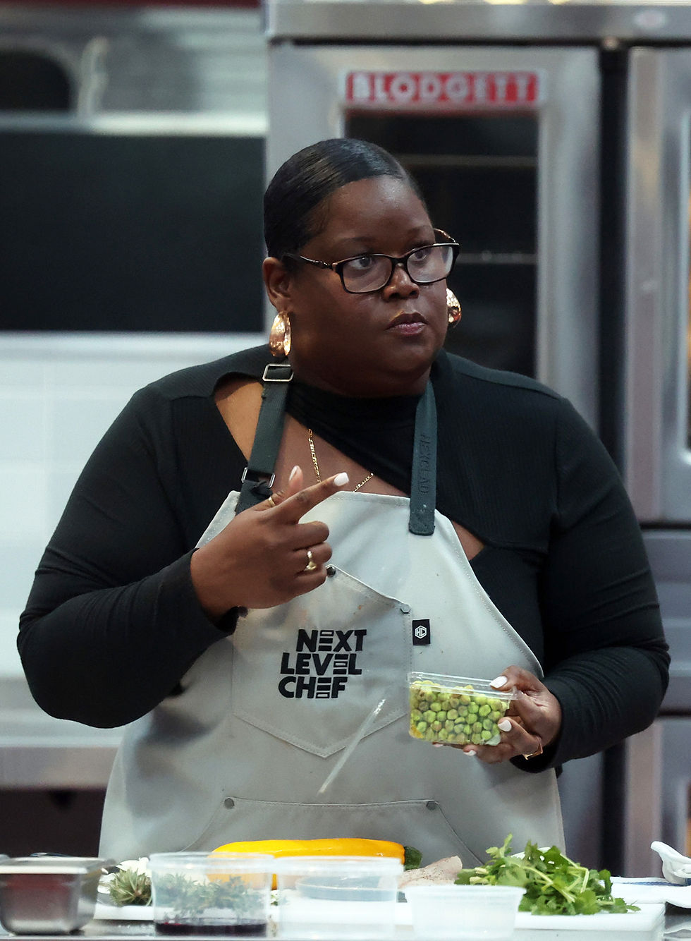 Chef in a kitchen wearing a "Next Level Chef" apron, holding a container of peas, with various ingredients on the counter. Focused expression.