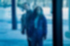 A man and woman in winter clothes stand outside in a snowy setting, holding papers and a book. They look serious, with a blue tint.