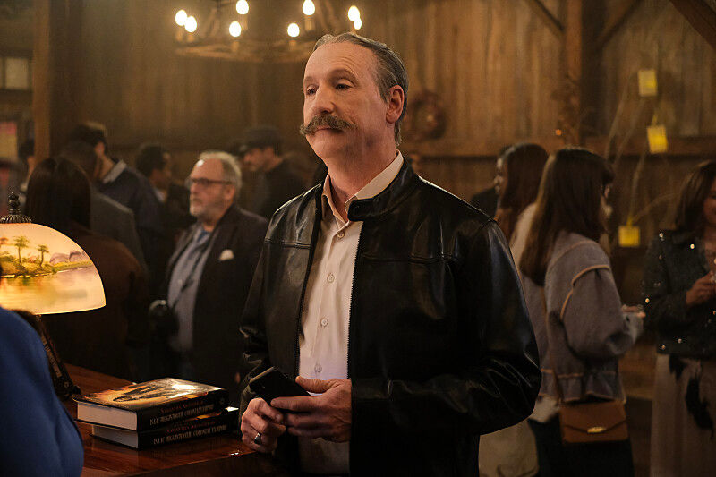 Man in leather jacket holds phone at a dimly lit gathering. Books and lamp with palm design on counter. Guests chat in rustic setting.