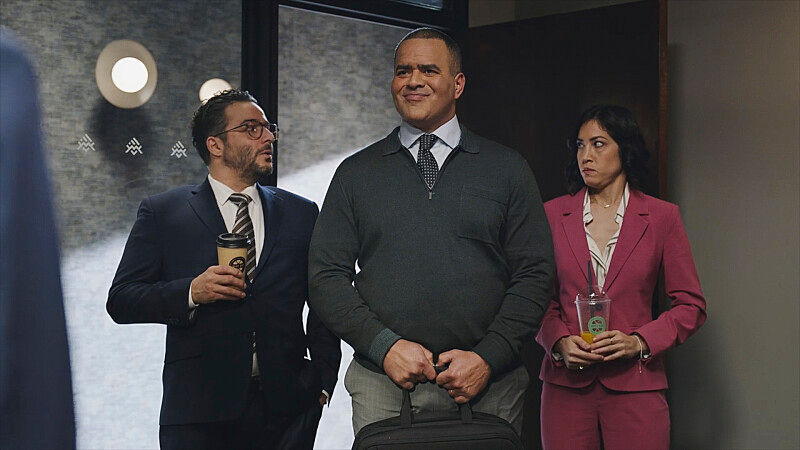 Two people in suits stand with another in a sweater, holding a bag. One holds coffee; the woman has a drink cup. Background shows lights.