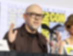 Bald man in glasses speaks into a microphone with hands raised at a Comic-Con panel, with blurred yellow event signage behind him.