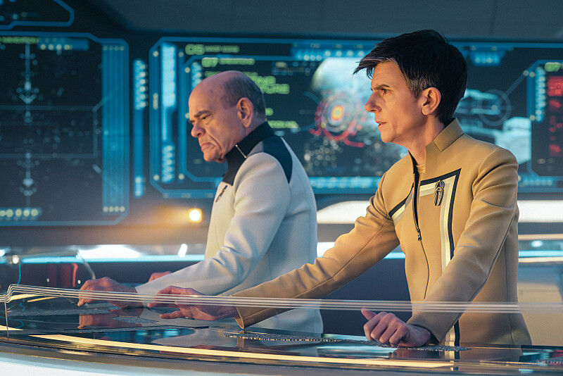 Two people in futuristic uniforms focus on glowing control panels with holographic displays. The room is bright with a tech-savvy ambiance.