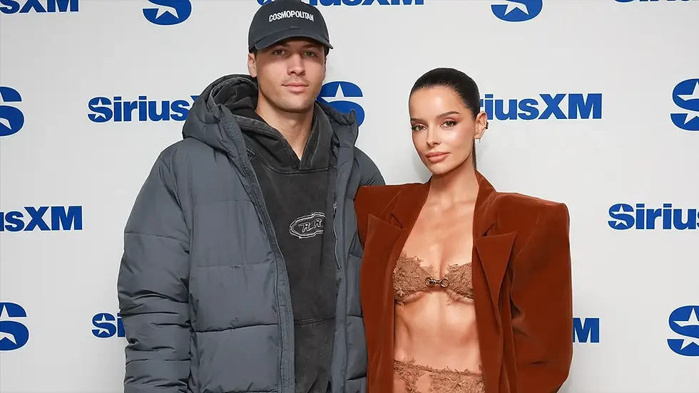 Two people pose at a SiriusXM backdrop; man in cap and gray jacket, woman in brown blazer and lace lingerie, both serious