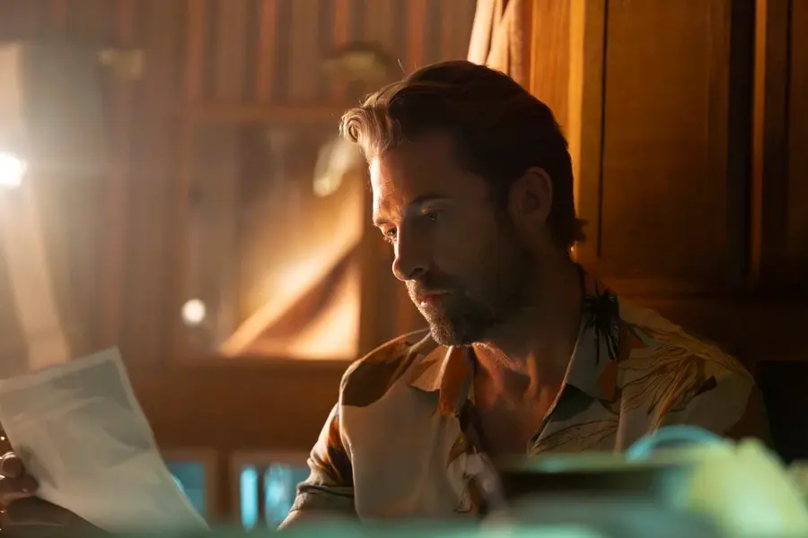 Man in a patterned shirt reads a document in a warmly lit room with wooden walls and a window in the background, creating a contemplative mood.