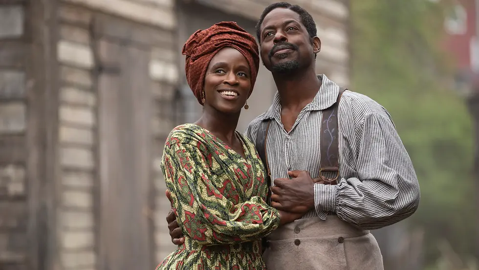 A smiling couple embraces outside a rustic wooden building. The woman wears a colorful patterned dress, the man is in striped suspenders, both look happy.