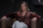 Long-haired man in a burgundy velvet shirt and striped pants sits in a leather chair in a dark studio, looking serious.