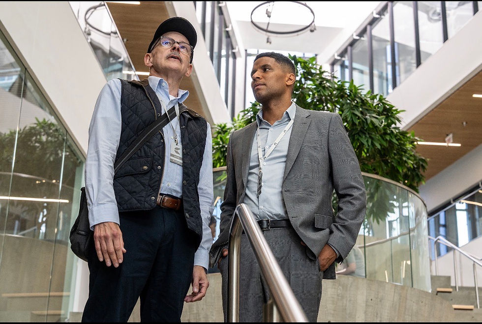 Two men in business attire stand on a staircase indoors. One wears a cap and vest, the other a gray suit. Modern building with plants.
