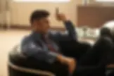 Serious man in a blue shirt and striped suit pants reclines in a black chair, holding a cigar in a bright office lounge.
