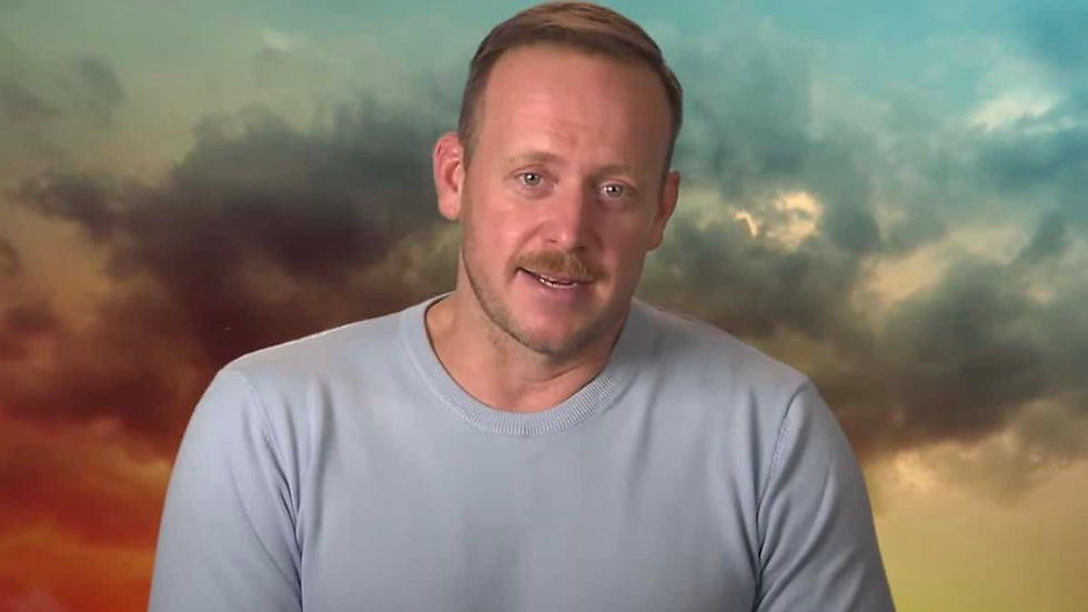 Man in a light blue shirt speaks to camera against a colorful cloudy sky backdrop.