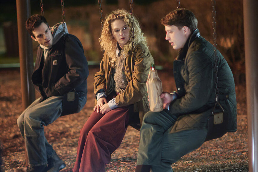 Three young adults sit on swings at night, talking quietly; one holds a bottle in a paper bag, with warm park lights behind.