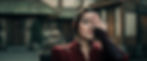 Woman in a red ornate robe covers one eye in a blurred courtyard, looking calm and serious.