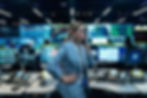 A woman in a blue suit speaks on the phone in a control room. Multiple monitors display maps and data, creating a focused atmosphere.
