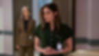 Two women in office attire stand in a modern hallway. One is in focus, appearing concerned, while the other looks on. Both wear ID badges.