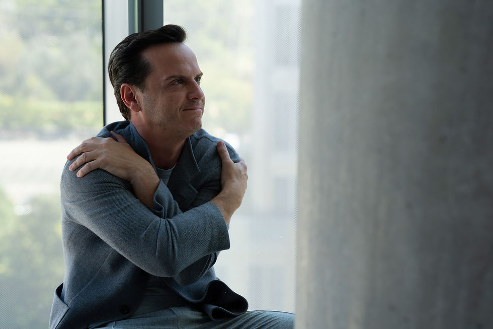 Man in gray suit embraces himself, sitting by a large window. Soft natural light, blurred greenery outside, calm and contemplative mood.