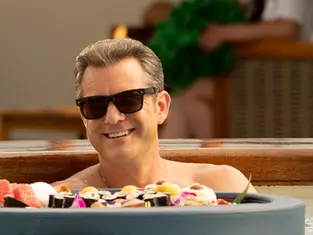 Man smiling in sunglasses, relaxing in a hot tub surrounded by sushi and colorful desserts. Background hints at a sunny outdoor setting.