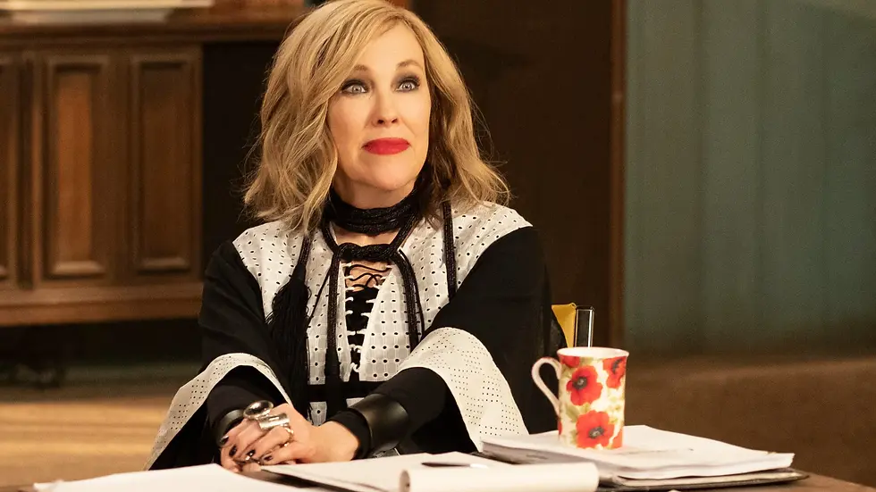 A woman with bold red lipstick and a patterned blouse sits at a desk with papers and a floral mug. She appears thoughtful indoors.