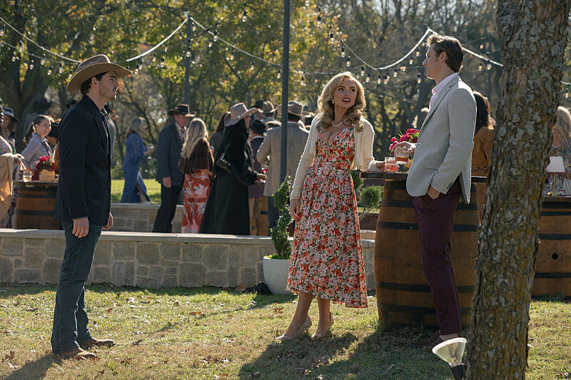 Cowboy-hatted man, floral-dress woman, and light-blazer man chat at a festive outdoor party with string lights and guests.