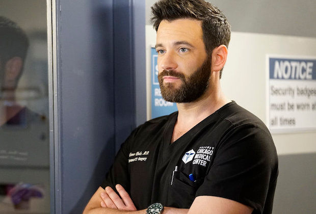Bearded man in black scrubs with Chicago Medical Center logo stands arms crossed in a hospital hallway, beside a notice sign.