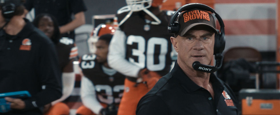 Cleveland Browns coach in headset and cap on sideline, looking tense, with blurred players and orange-black gear behind him.