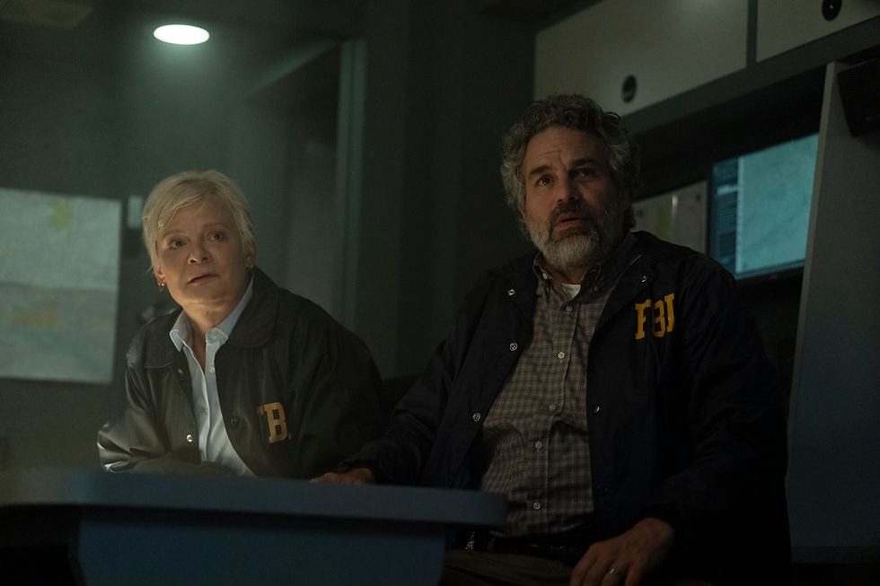 Two people in dark FBI jackets sit at a desk in a dim room, looking intently at something. Monitors and maps are visible in the background.