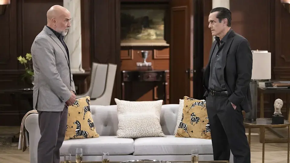 Two men stand facing each other in a formal room, looking tense. A white sofa with patterned pillows is between them. Dark wood decor.