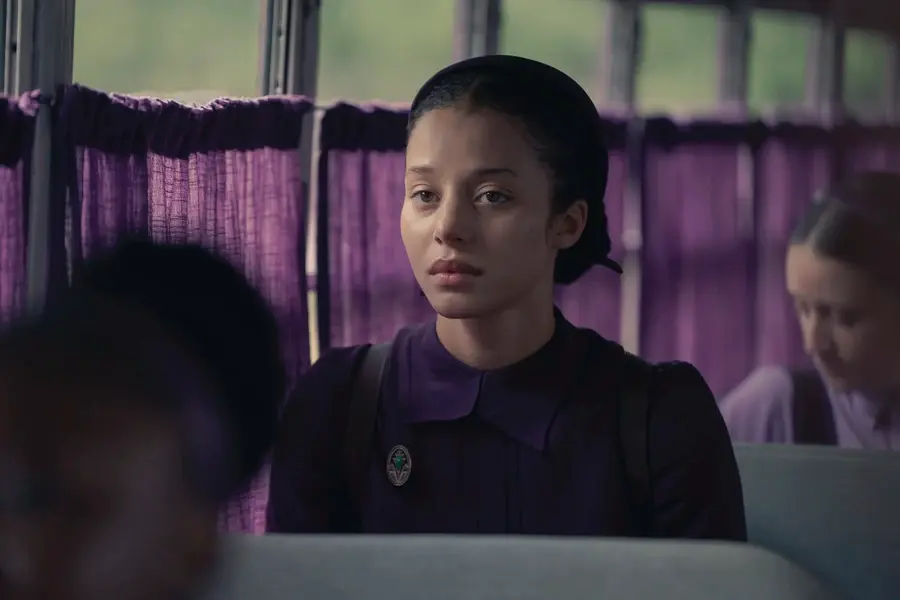 Serious young woman in a dark purple uniform sits on a bus, with purple curtains and blurred passengers behind her.