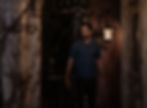 Man in a blue polo stands on a dimly lit porch, surrounded by vines and aged wood. A lantern casts a warm glow, creating a mysterious mood.