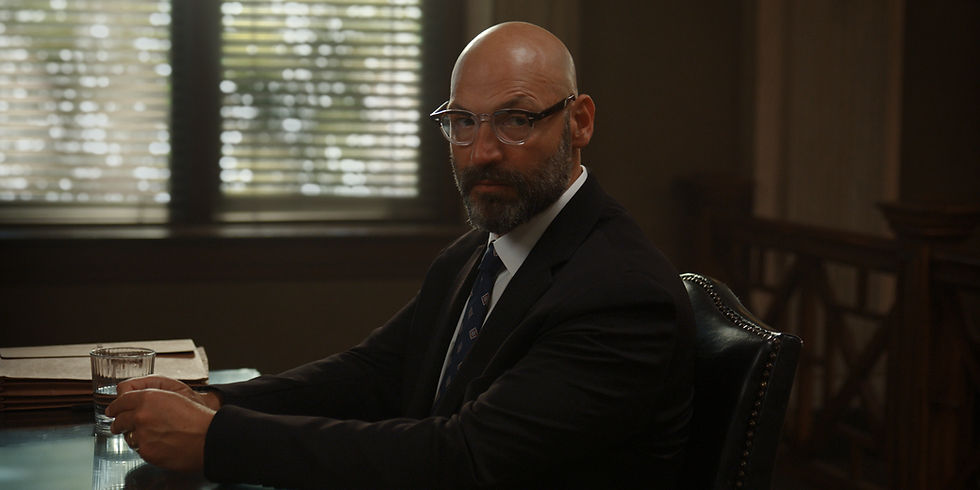 Bald man with glasses and suit sits at a wooden table holding a glass of water. Background has blurred window blinds with soft light.