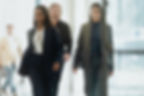 Three business professionals walk through a bright office corridor, with two women in sharp suits leading and a man behind.