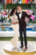 Smiling couple pose arm in arm on a lit stage in formal outfits, with colorful lights and a pool behind them, celebrating.