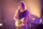 Long-haired guitarist in a black tank top plays a white electric guitar on a purple-lit stage, with drums behind him and a focused stare.