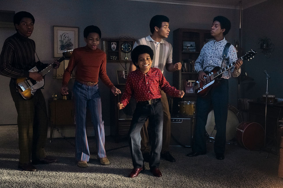 Five young musicians perform energetically indoors. Two play guitars. Vibrant 70s attire and a cozy living room create a lively atmosphere.