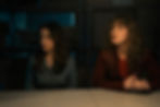 Two women sit at a table in a dimly lit room with shelves. They look attentive and concerned. The mood is tense and focused.