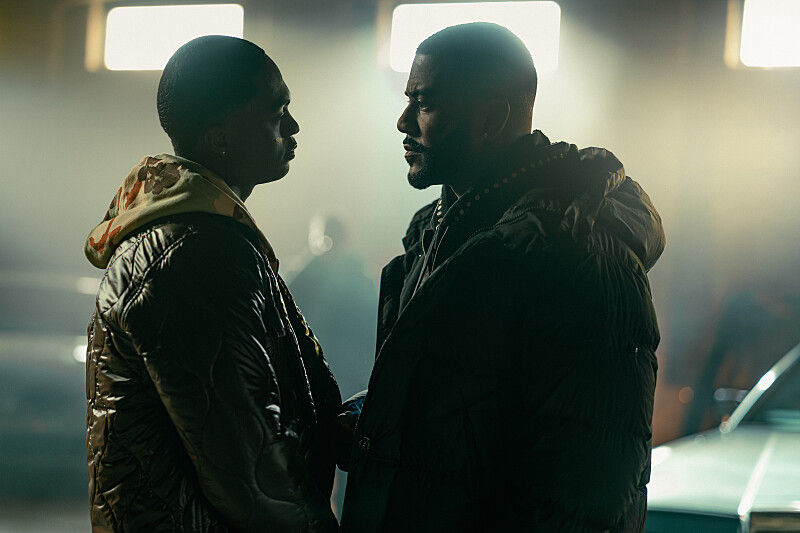 Two men in dark puffer jackets stand face to face in a dim garage, lit by bright lights, with a tense mood.