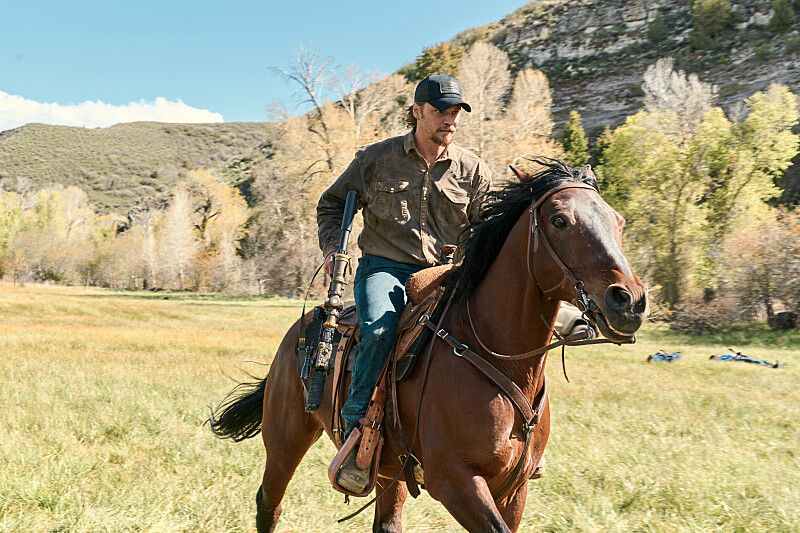 Man rides a brown horse through a grassy meadow, rifle in saddle, with mountains and autumn trees behind.