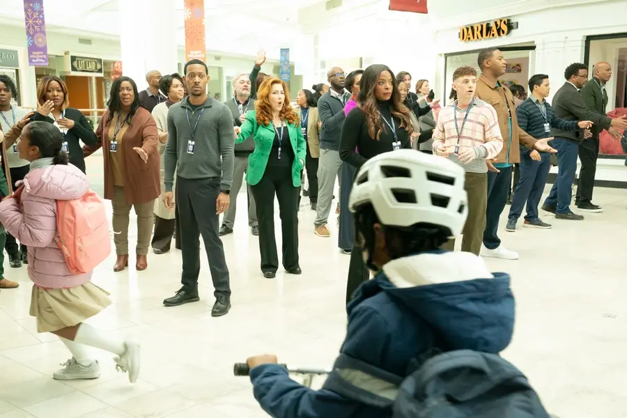 Crowd of people in a mall look upward, surprised. A child with a pink backpack and a cyclist in a helmet pass by. Bright banners above.