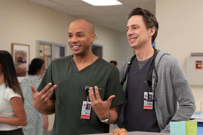 Two smiling doctors in a hospital hallway, one in green scrubs gesturing and one in a gray cardigan with a stethoscope.