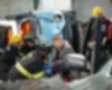 Firefighters in helmets assist a man near a flipped, damaged blue vehicle. The scene is intense and focused, suggesting an emergency.