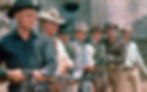 Seven cowboys in hats and various outfits stand in a line outdoors, holding rifles, with a tense, focused mood. Stone wall in background.