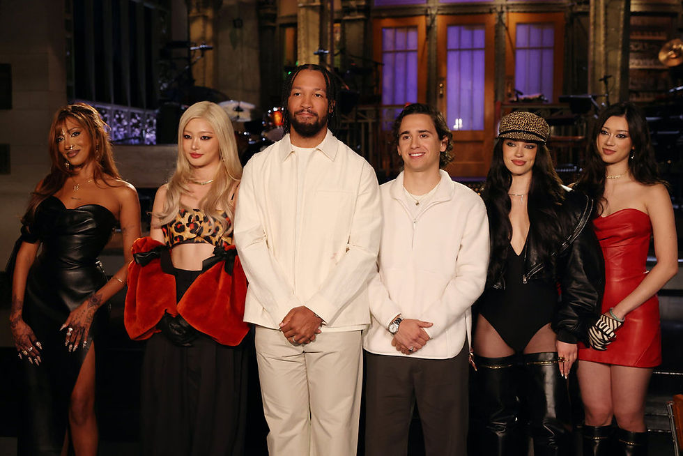 Five performers pose on a lit studio stage; two men in white center, three women in black, red, and leopard print outfits.