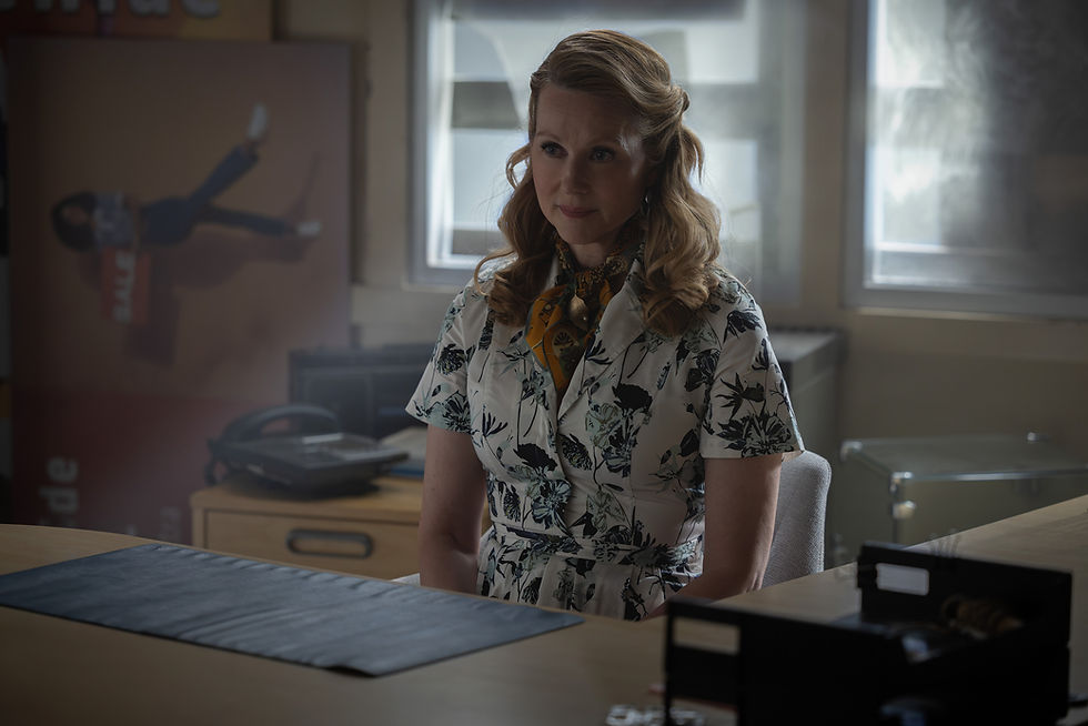 Woman in floral dress sits in a dim office, looking thoughtful; windows glow behind her and a blurred poster hangs nearby.