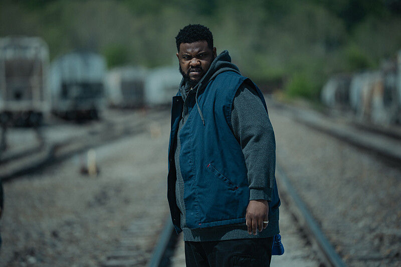 Man in a blue vest stands on train tracks, looking intensely at the camera. Blurry trains in the background, overcast mood.