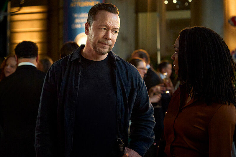 Man in dark jacket speaks with a woman in a brown top at a dim indoor event, with a blurred crowd and warm lights behind them