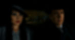 Two people in a car at night, wearing vintage attire. The woman sports blue eyeshadow and red lipstick. The man wears a suit and hat.