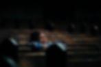 Person in a blue jacket sits alone on a wooden bench in a dimly lit space, eyes closed, appearing contemplative or emotional.